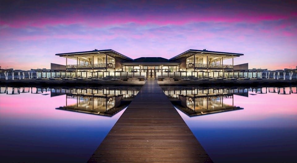 Gebouw met veel ramen en een houten steiger aan het water met roze lucht
