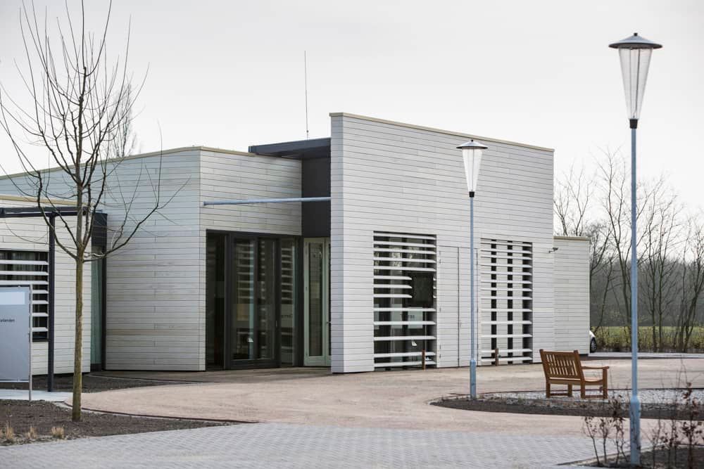 Wit houten gebouw met ramen met witte strepen ervoor en een pleintje met een bank, lantaarnpalen en bomen