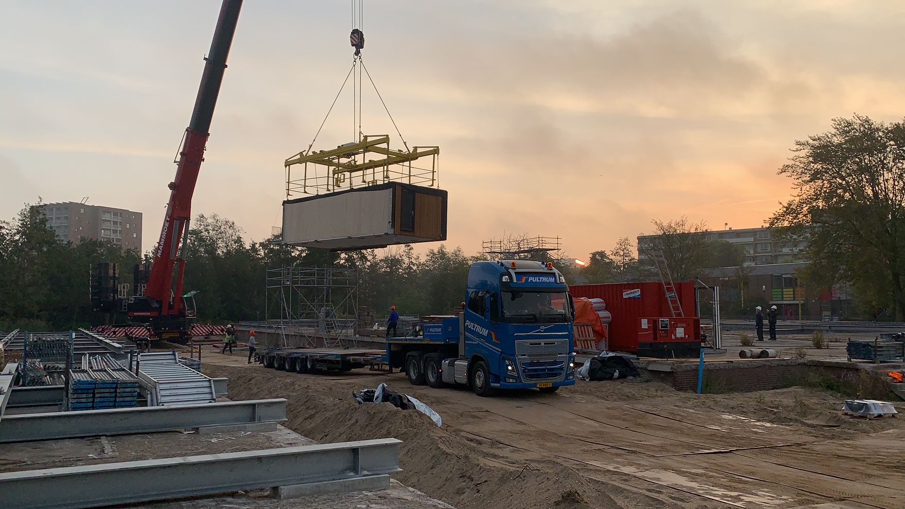 Bouwplaats van zand met houten module aan een hijskraan met een vrachtwagen eronder