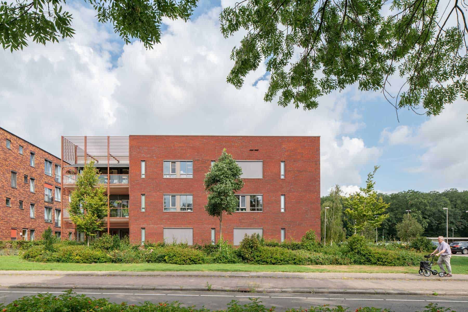 Het rood bakstenen woonzorgcomplex vanaf de straatkant met vooraan een man met rollator