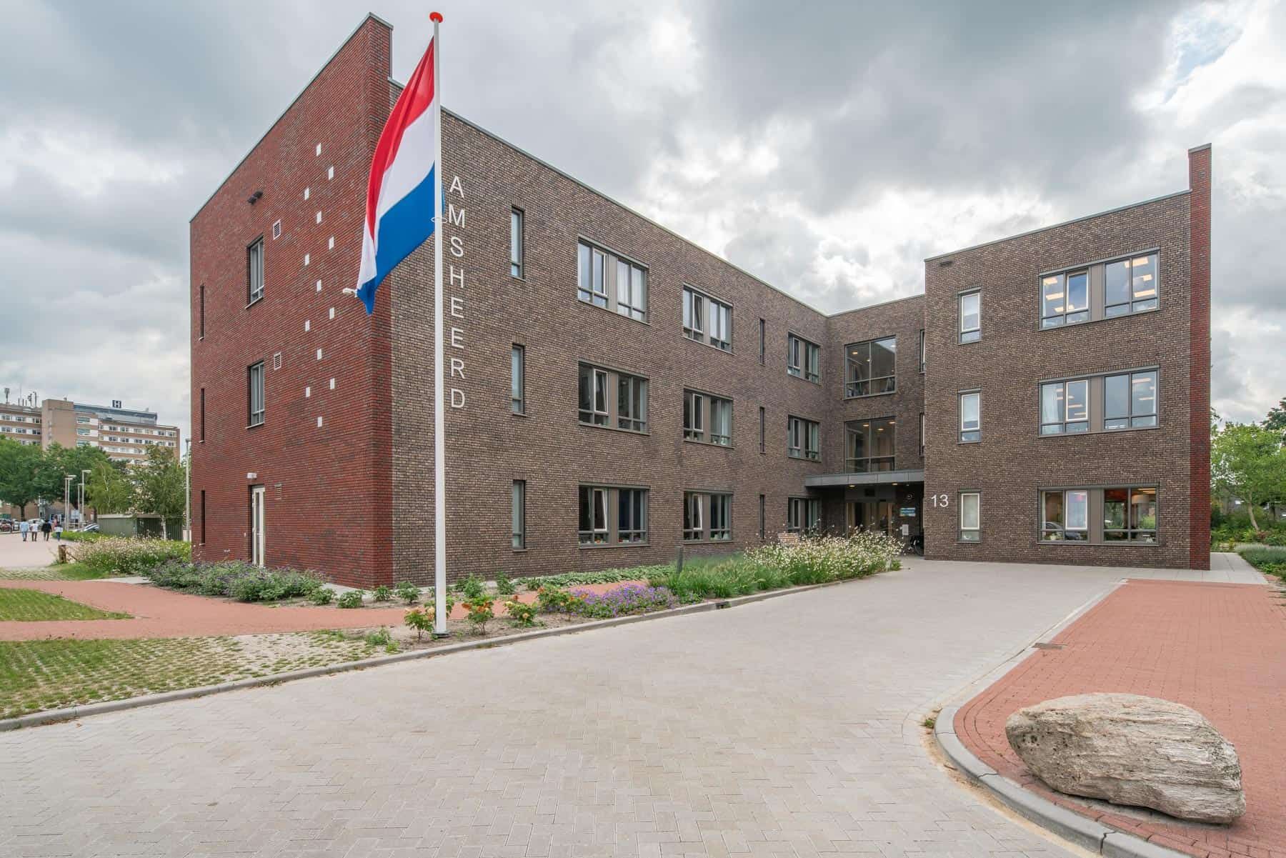 De oprijlaan van woonzorgcomplex met de bruine bakstenen aan de voorkant, rode aan de zijkant en een Nederlandse vlag