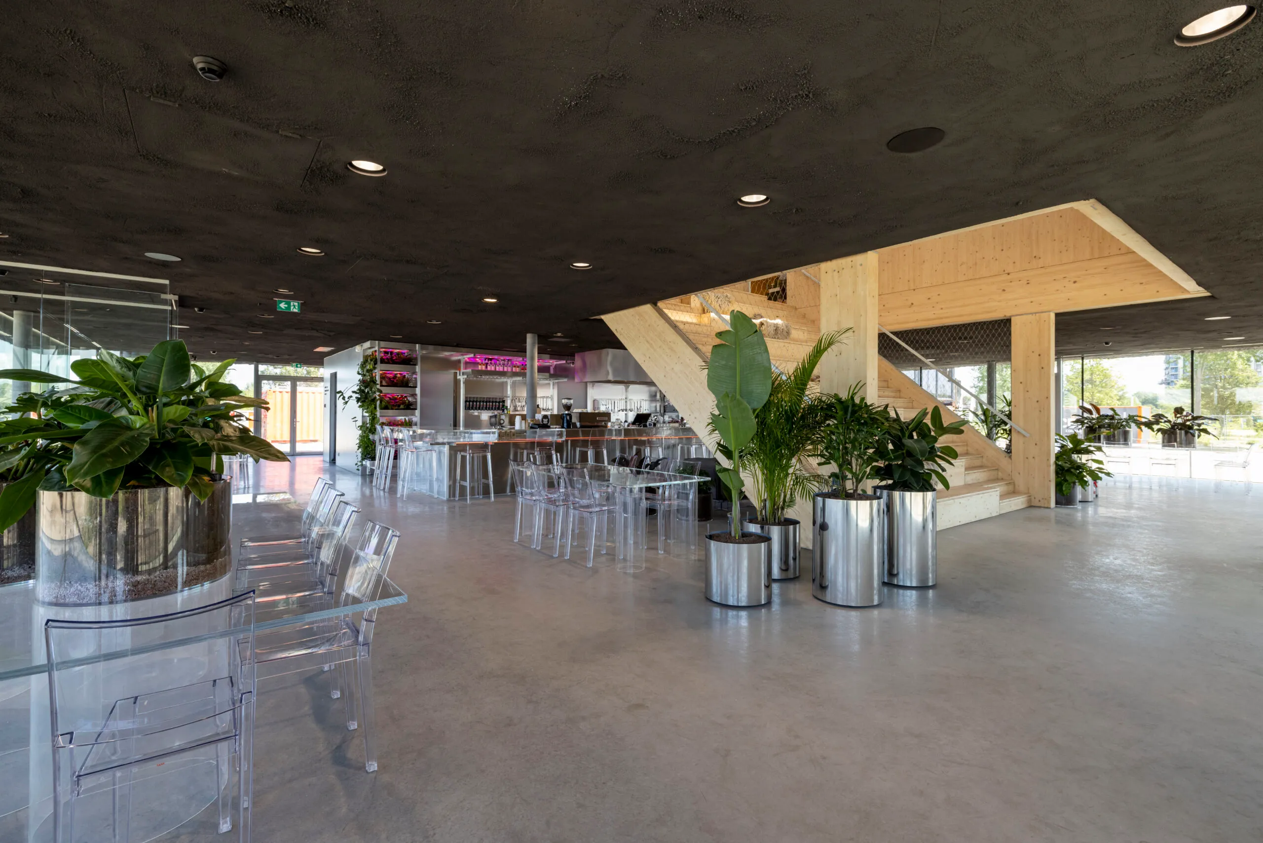 Restaurant met doorzichtig meubilair, veel planten, een bar en een houten trap