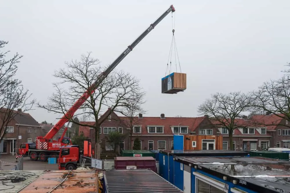 Een lichtbruine box die wordt vervoerd via een rode, hoge kraan