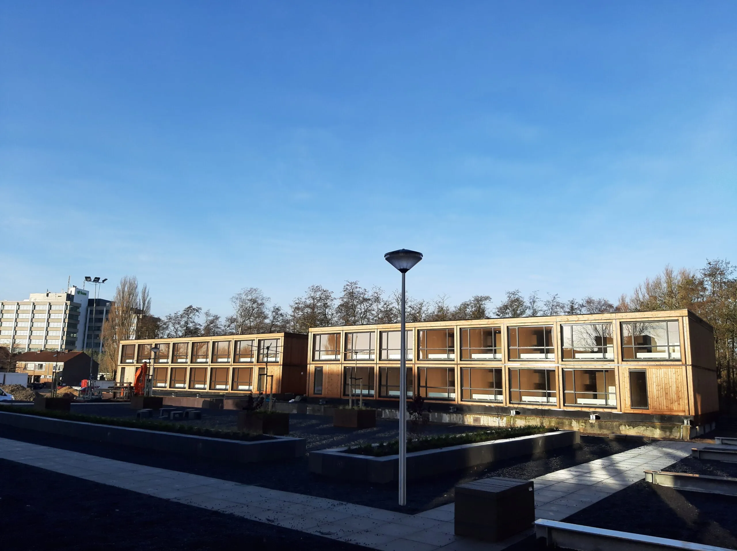 Houten gebouw met grote ramen voor bomen en grind, een pad van stoeptegels en een lantaarnpaal ervoor