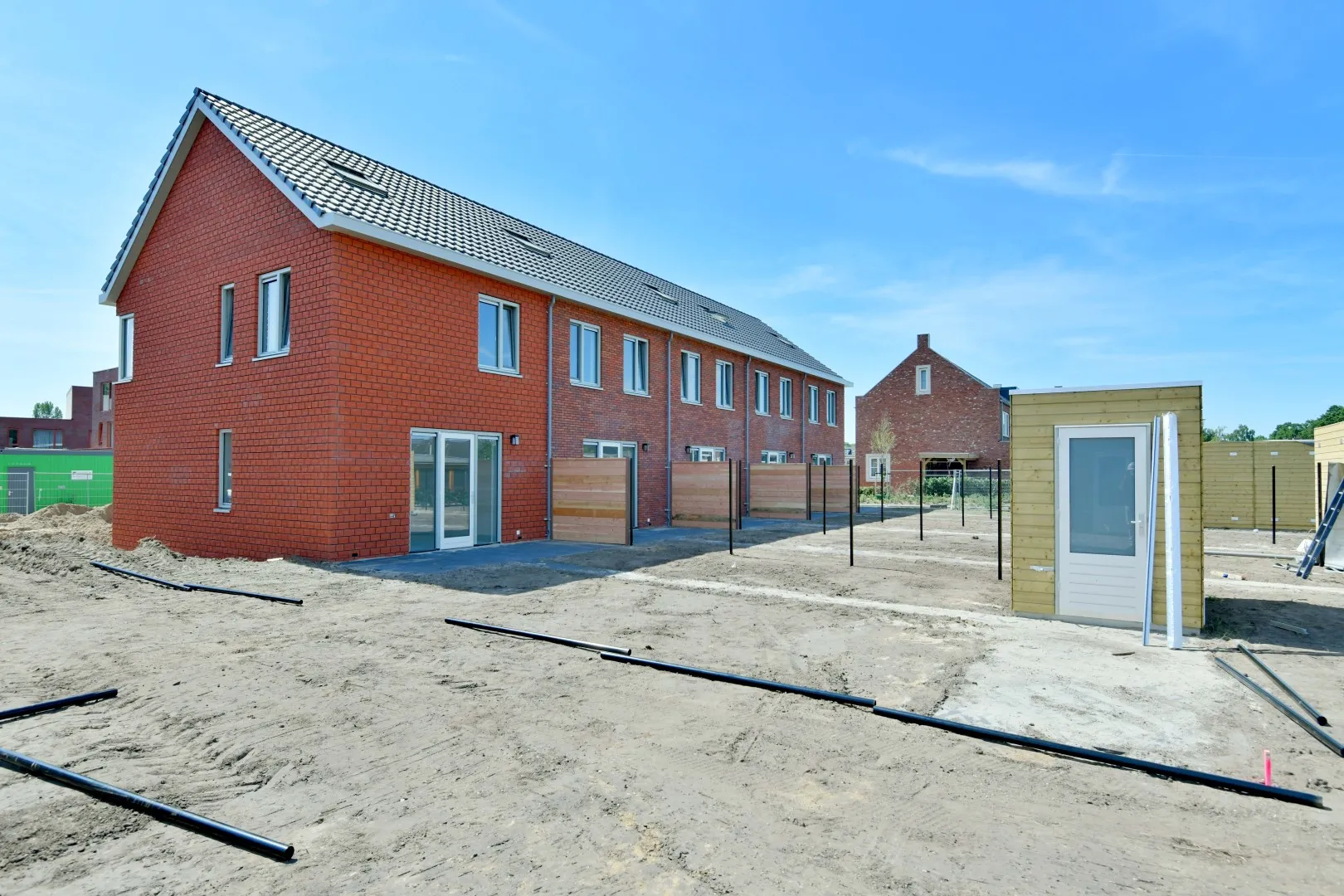 Rode nieuwbouw rijtjeshuizen met zwarte daken met gele, houten berging in tuin.