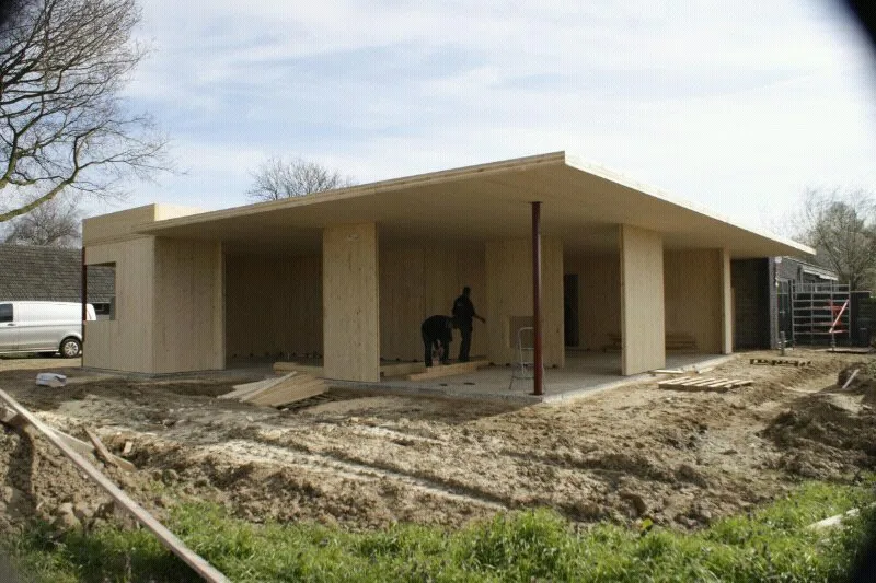 Houten gebouw op zand en gras waar twee mensen aan het werk zijn
