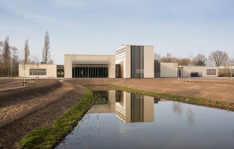 Witte vierkante gebouwen met zwarte strepen achter een vijvertje en zandvlakte