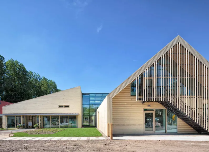 Gebouw van hout met links een schuin dak, rechts glas en daarnaast een puntdak met een trap aan de buitenkant met balkjes naar beneden ervoor